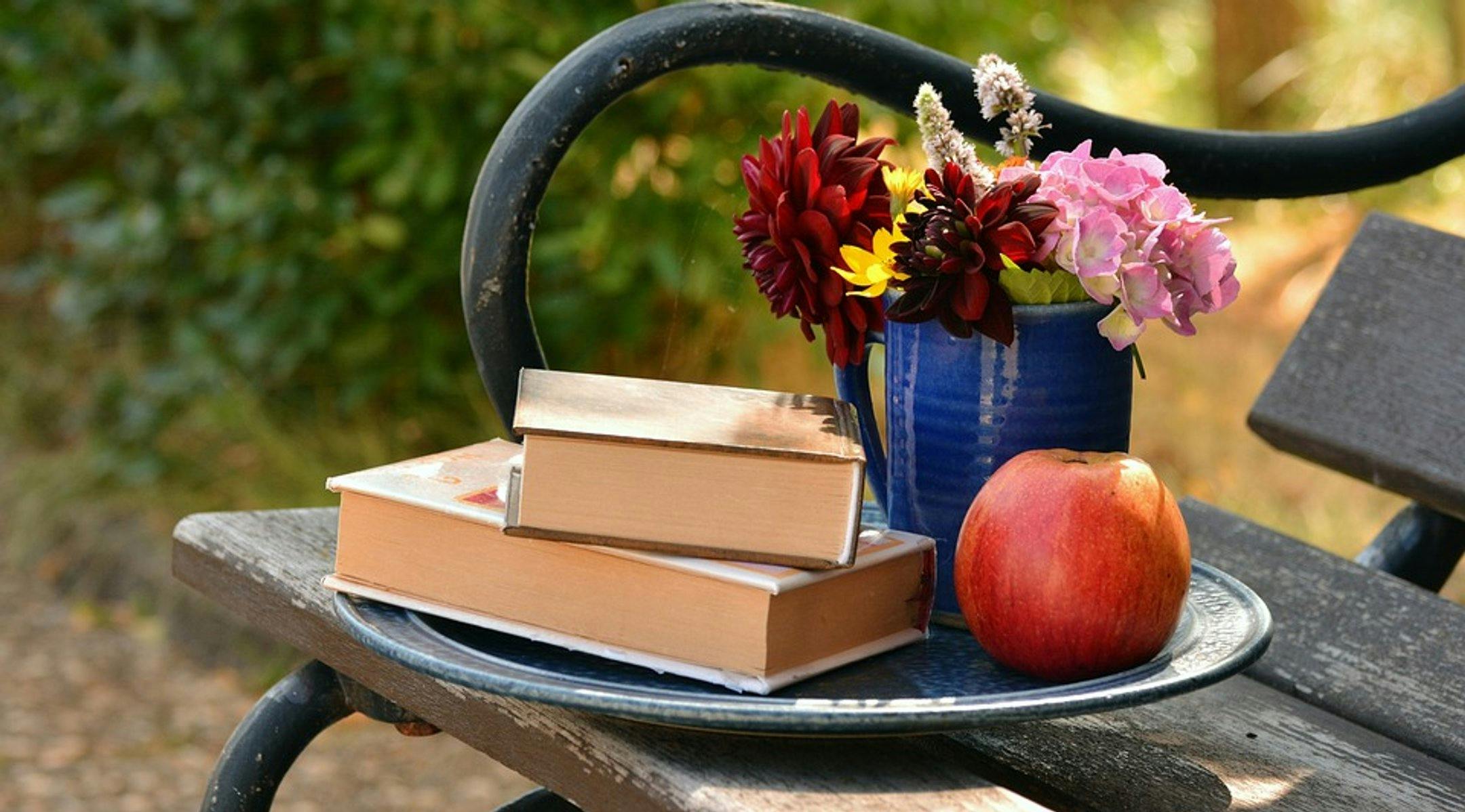 En benk med et fat oppå. På fatet ligger to bøker, et eple og blomster i et krus.
